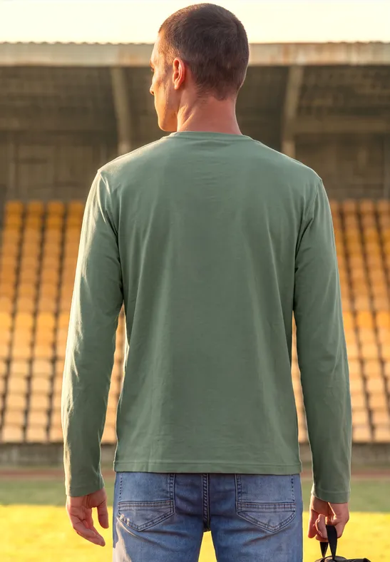 Ein Mann in Jeans und grünem Shirt steht im Stadion und hält einen roten Football-Helm.