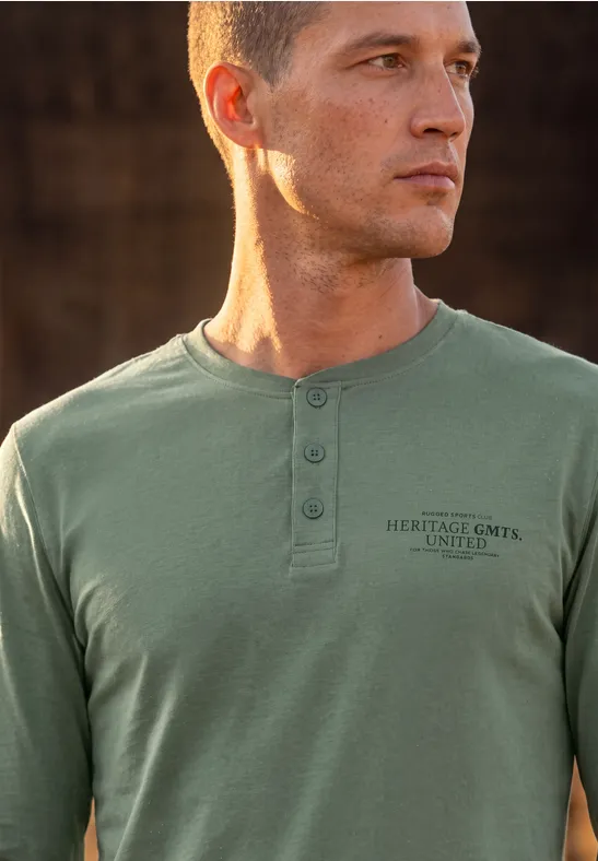 Ein junger Mann mit kurzem Haar trägt ein olivgrünes Langarmshirt mit Knopfleiste und Print.