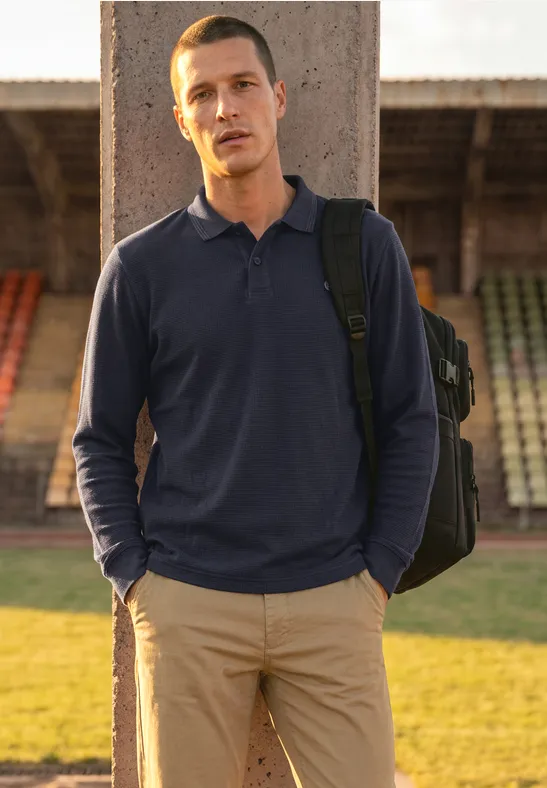 Ein Mann mit Rucksack steht vor Stadiontribüne, trägt dunkelblaues Polo und helle Hose.