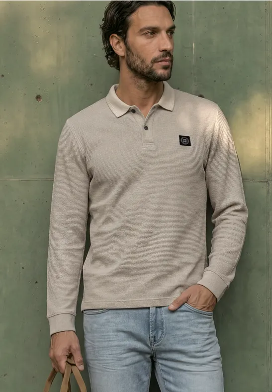 Ein bärtiger Mann in beiger Langarm-Poloshirt und Jeans lehnt vor einer grünen Wand.