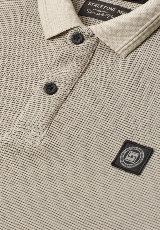 Nahaufnahme eines beigen Pikee-Poloshirts mit schwarzem Logo-Patch auf der Brust und zwei Knöpfen.