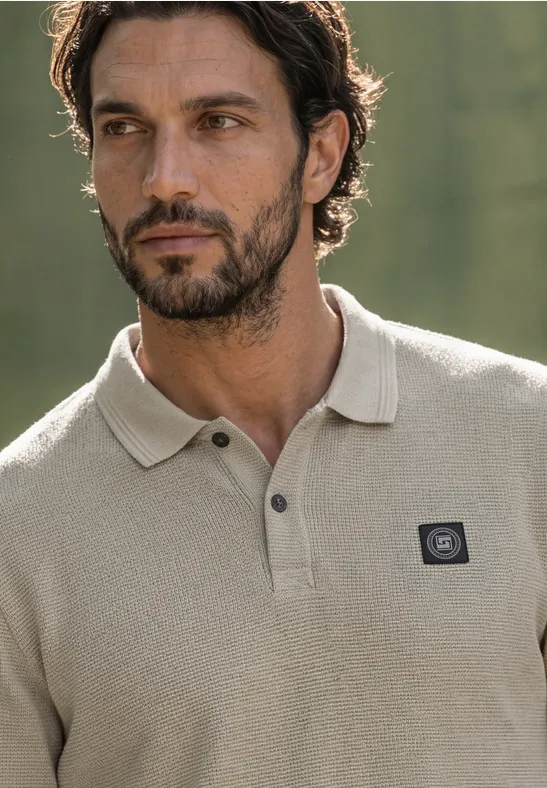 Ein Mann mit leichtem Bart trägt ein beiges Poloshirt vor unscharfem Naturhintergrund.