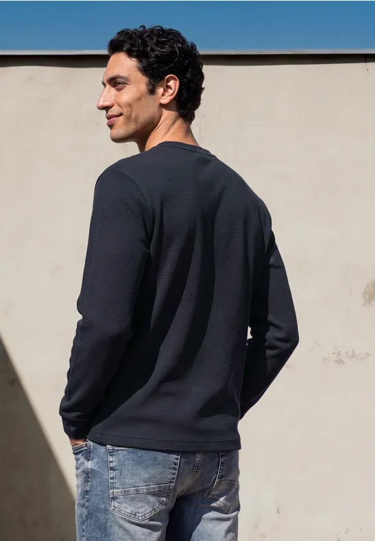 Ein junger Mann steht seitlich, trägt schwarzen Pullover und Jeans vor einer beigen Wand.
