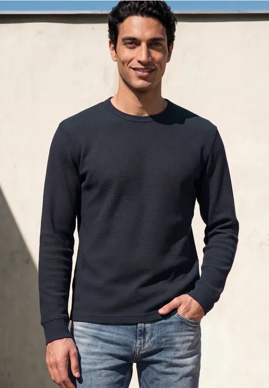 Ein lächelnder Mann in schwarzem Pullover und Jeans steht vor heller Wand im Freien.