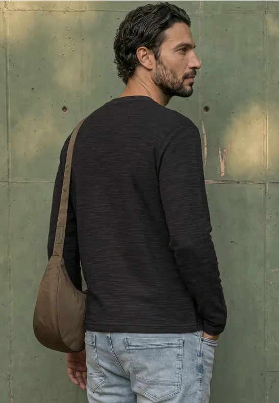 Ein bärtiger Mann in schwarzem Pullover und hellen Jeans trägt eine grüne Schultertasche vor einer grünen Wand.
