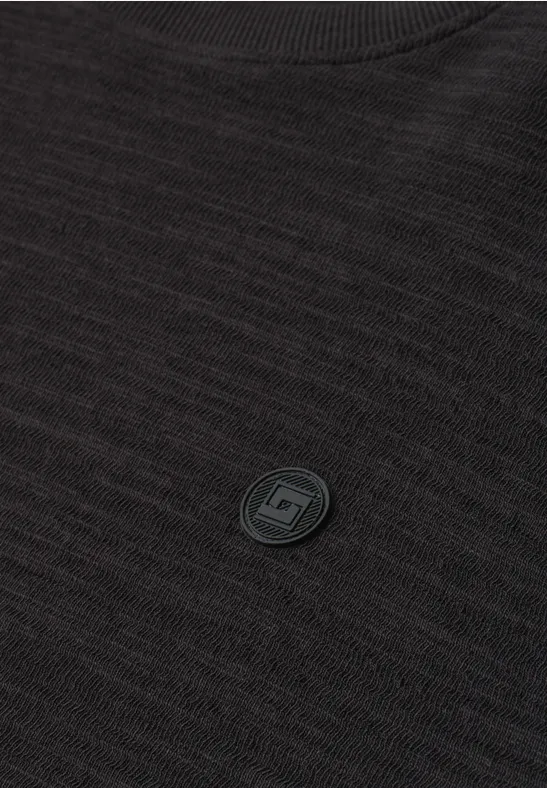 Nahaufnahme eines schwarzen Strickpullovers mit rundem Ausschnitt und kleinem, rundem Logo-Emblem auf der Brust.