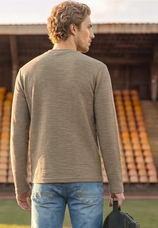 Ein Mann steht von hinten vor Tribüne, trägt grünen Pullover, Jeans und hält eine Tasche.