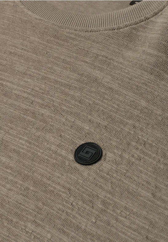 Nahaufnahme eines braunen Strickshirts mit feiner Struktur und schwarzem Rundlogo auf der Brust.
