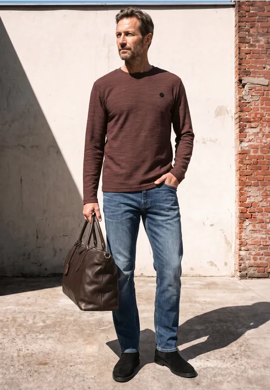 Mann im braunen Pullover, blauen Jeans und schwarzen Schuhen trägt eine große Ledertasche, steht im Sonnenlicht.