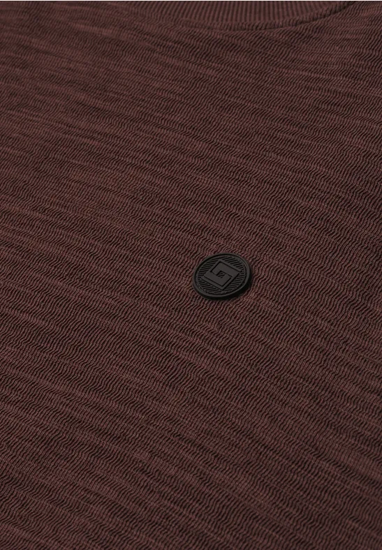Nahaufnahme eines braunen Strickpullovers mit schwarzem, rundem Logo-Badge