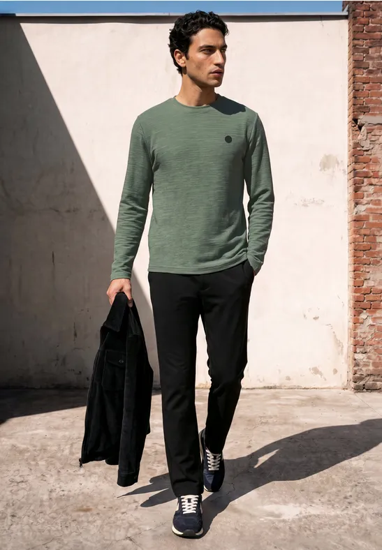 Ein Mann in grünem Pullover und schwarzer Hose hält eine Jacke und geht draußen in urbaner Umgebung.