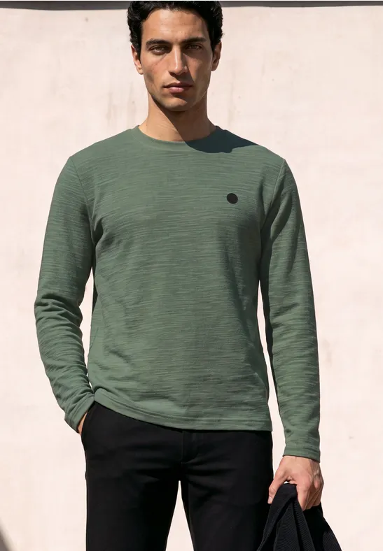 Ein junger Mann vor einer rosa Wand, trägt ein grünes Langarmshirt, schwarze Hose und hält eine schwarze Tasche.