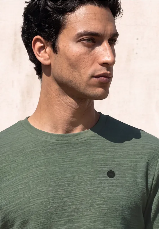 Ein junger Mann mit dunklen Haaren in einem grünen Shirt blickt seitlich vor hellem Hintergrund.