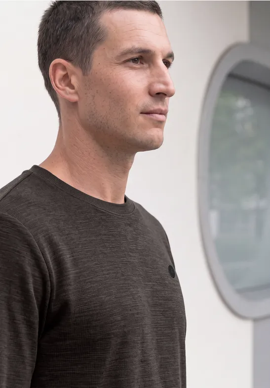 Ein Mann mit kurzem Haar und dunklem T-Shirt blickt seitlich in eine helle, moderne Umgebung.