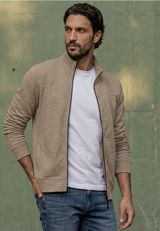 Ein Mann in beiger Strickjacke, weißem T-Shirt und Jeans vor grünem Hintergrund.