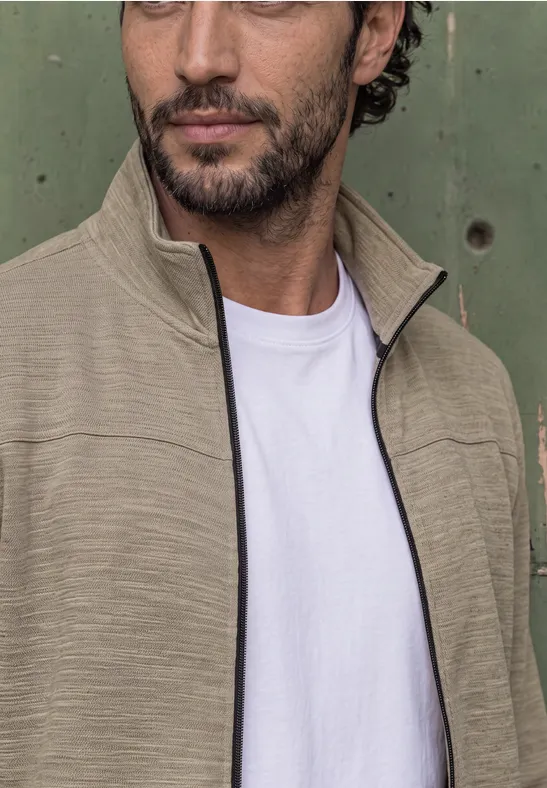 Ein Mann mit dunklen Haaren und Bart trägt eine beige Jacke über weißem Shirt vor grünem Hintergrund.
