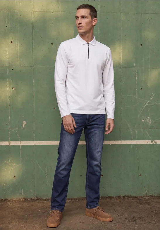 Ein Mann in weißem Langarmshirt, blauen Jeans und braunen Schuhen steht vor einer grünen Wand.