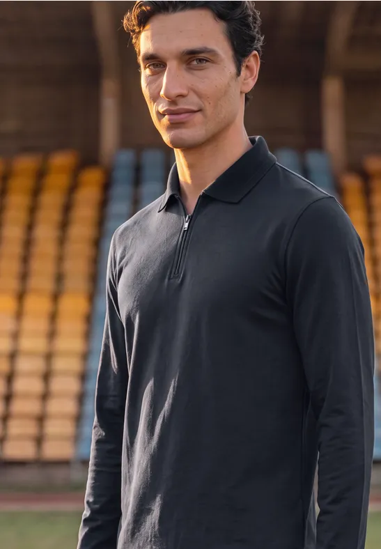 Ein sportlich gekleideter Mann in schwarzem Langarm-Polo steht vor Stadiontribünen.