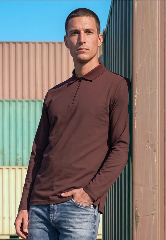 Ein Mann in braunem Poloshirt und Jeans lehnt vor bunten Containern und blauem Himmel.