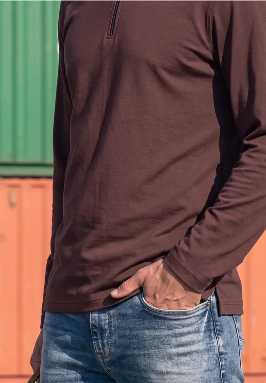Ein Mann trägt ein braunes Langarmshirt und Jeans vor grünen und orangenen Containern.