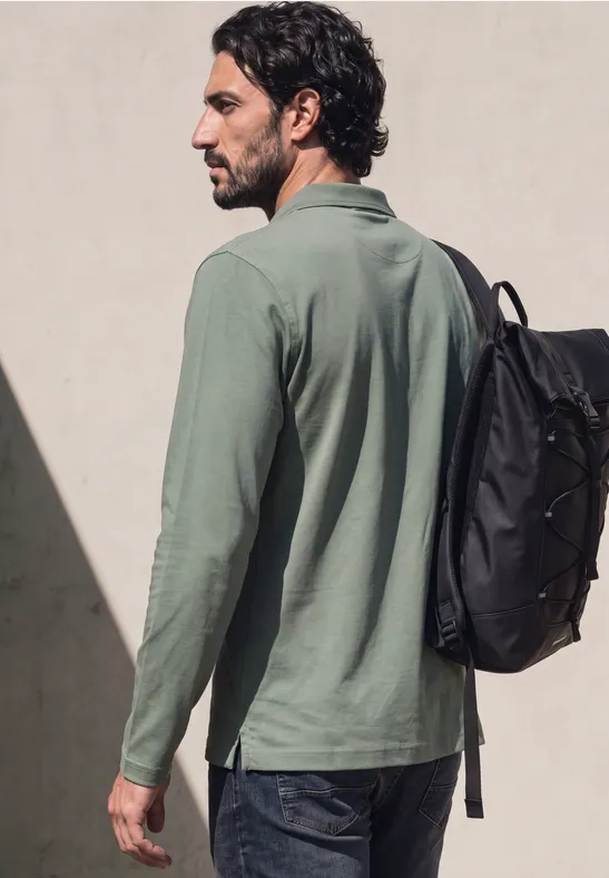 Ein Mann mit grünem Langarmshirt und Rucksack schaut seitlich vor einer hellen Wand.