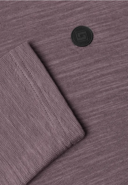 Violetter Strickstoff mit sichtbarer Maschenstruktur, umgeschlagener Ecke und rundem schwarzen Logo.