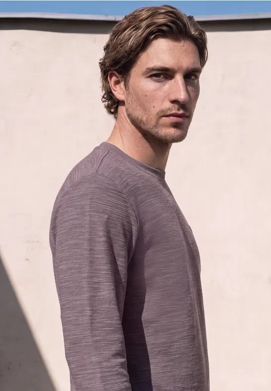 Ein Mann mit braunen Haaren trägt einen grauen Pullover vor einer hellen Mauer.