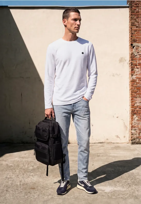 Ein Mann in weißem Langarmshirt und Jeans trägt einen schwarzen Rucksack und steht vor einer hellen Mauer.