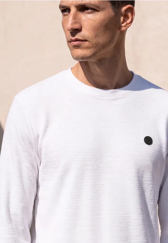 Ein Mann trägt ein schlichtes weißes T-Shirt mit einem kleinen schwarzen Punkt auf der Brust.
