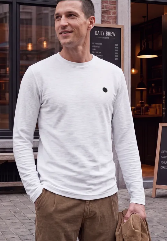 Een man in een wit lang mouw T-shirt en een bruine broek staat voor een café en glimlacht.