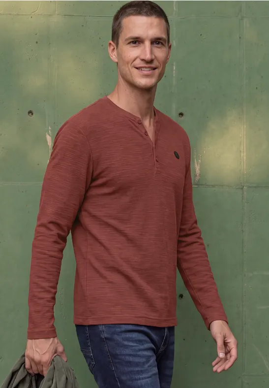 Un homme en t-shirt à manches longues rouge et jean se tient devant un mur vert, quelque chose à la main.