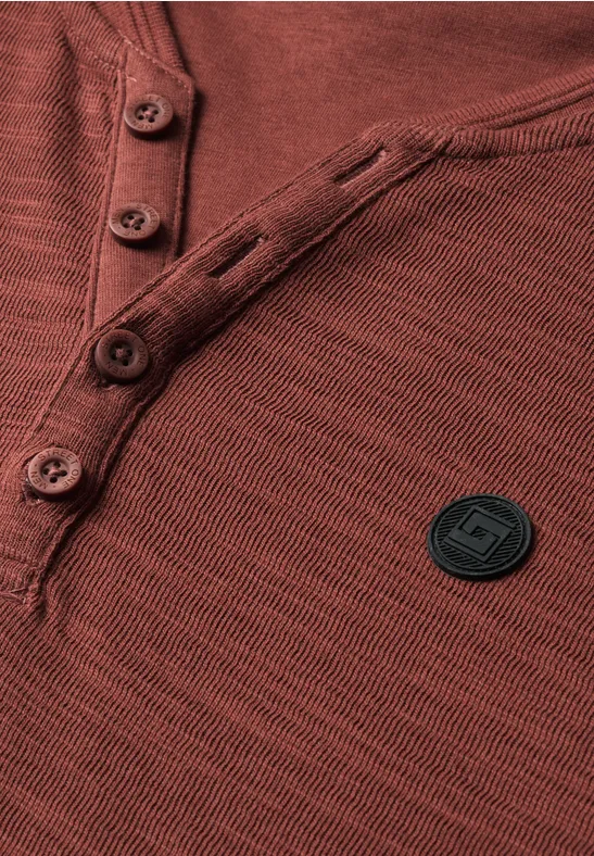 Gros plan d'un t-shirt Henley rouille avec patte de boutonnage et patch logo rond noir.