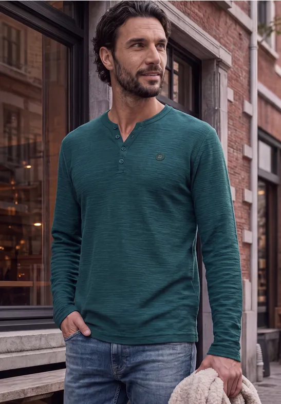 Un homme en t-shirt vert à manches longues et jean pose décontracté dans la rue.