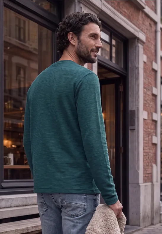 Un homme en t-shirt vert à manches longues et jean marche devant un bâtiment tout en regardant par-dessus son épaule.
