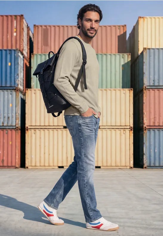 Ein Mann mit Rucksack, Jeans und Sneakern steht seitlich vor bunten Frachtcontainern.