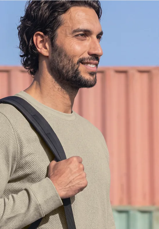 Ein lächelnder junger Mann mit dunklen Haaren trägt seinen Rucksack vor buntem Containerhintergrund.
