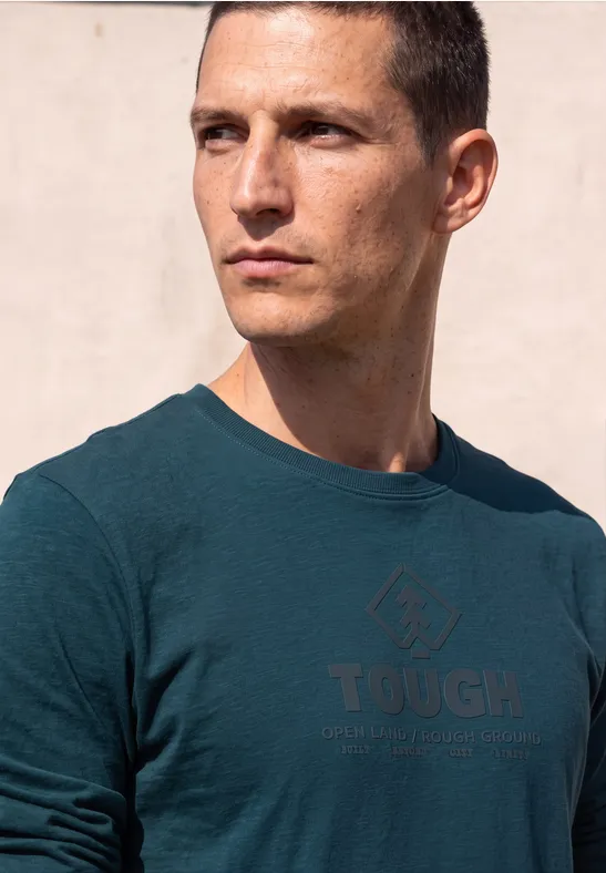 Mann mit kurzem Haar in grünem T-Shirt mit 'TOUGH'-Schriftzug blickt nach rechts.