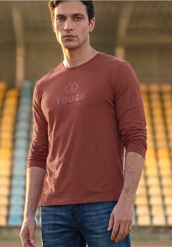 Mann in braunem Langarmshirt mit Aufschrift "TOUGH" vor Tribünen.