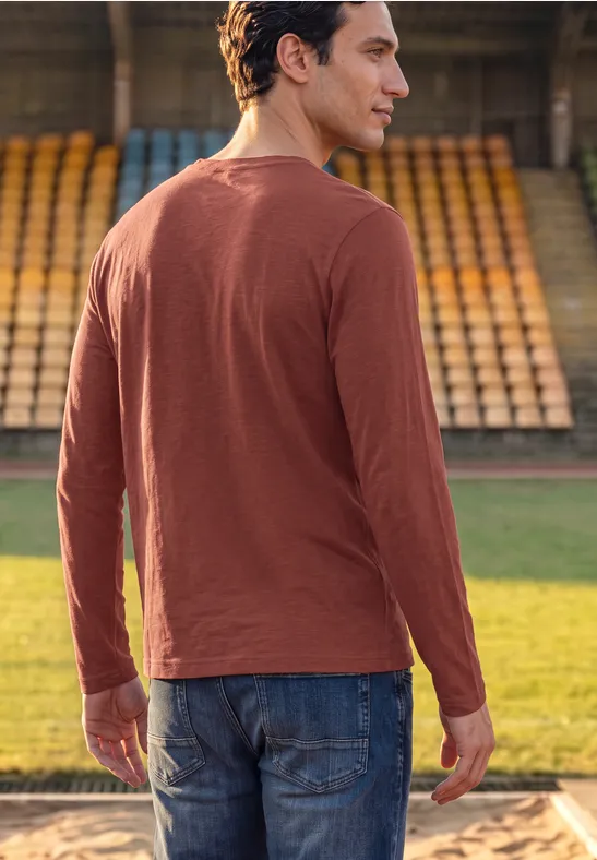 Ein Mann blickt von hinten auf das Spielfeld, trägt ein rostfarbenes Langarmshirt und Jeans.