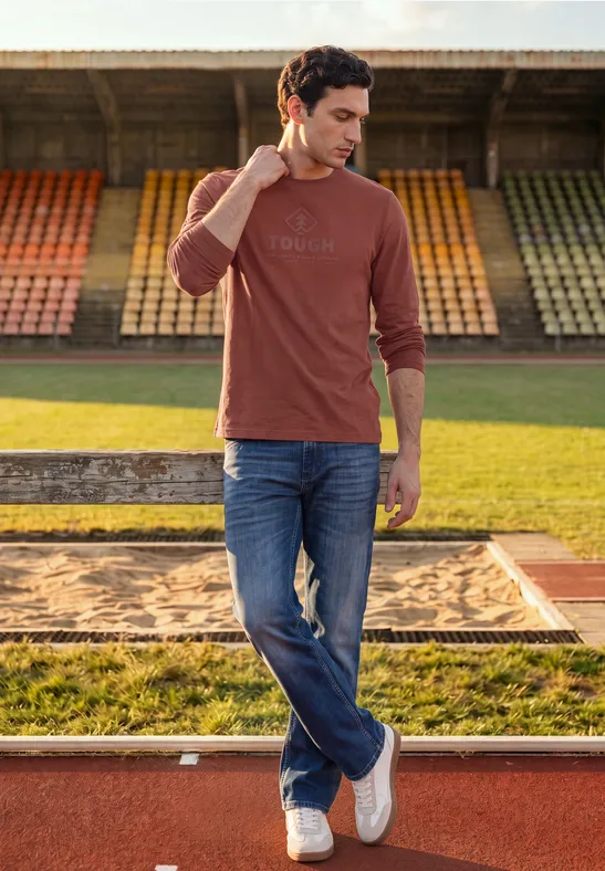 Ein Mann in Jeans und langärmeligem T-Shirt steht entspannt vor Stadiontribüne.