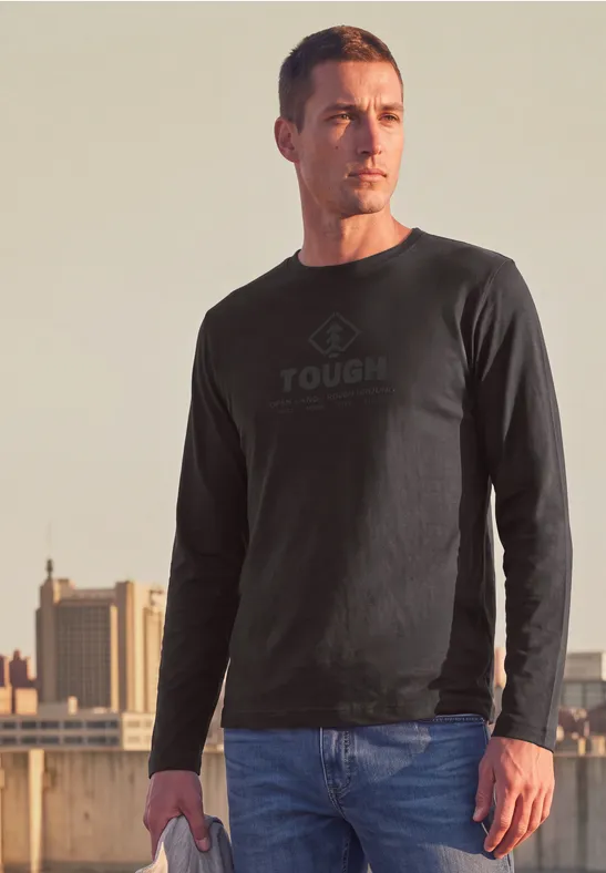 Un homme porte un sweat-shirt noir marqué 'TOUGH' sur un toit, avec une silhouette urbaine en arrière-plan.
