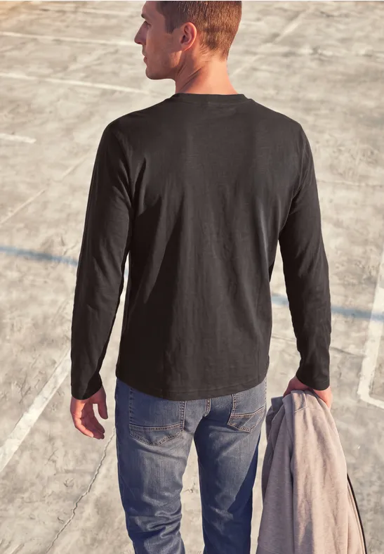 Un homme de dos en T-shirt noir à manches longues et jean, sur un terrain en béton, tenant un manteau.