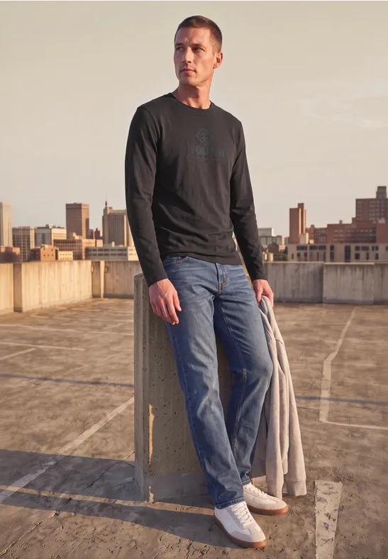 Homme sur un parking sur toit, pull noir, jeans, manteau à la main, skyline en arrière-plan.