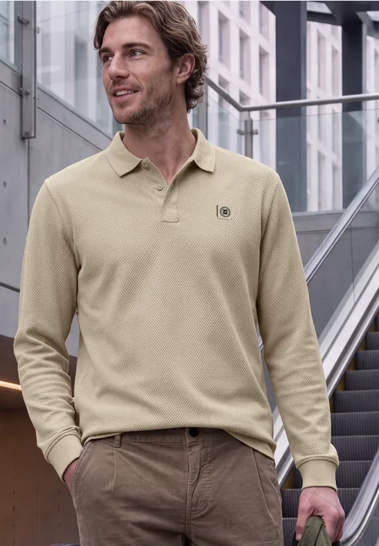 Mann in beigem Langarm-Poloshirt und brauner Hose, lässig stehend vor moderner Treppe.