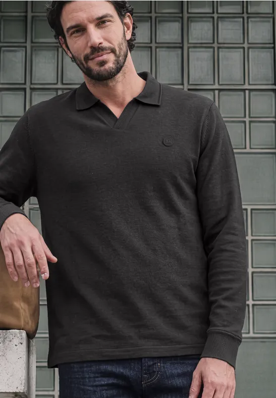Un homme appuyé sur un mur de briques de verre porte un polo noir à manches longues et un jean.