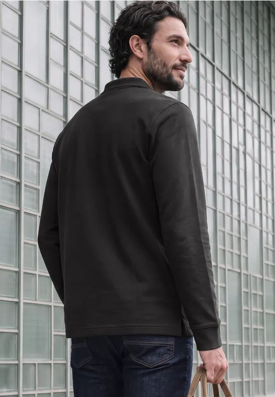 Un homme barbu en chemise noire et jeans est debout devant un mur en verre et regarde sur sa droite.