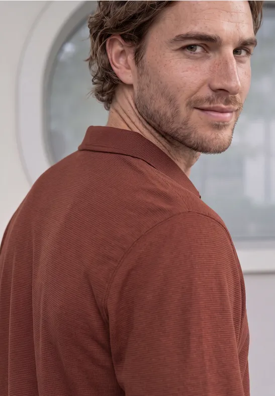 Ein leicht lächelnder Mann im braunen Poloshirt blickt seitlich über die Schulter vor hellem Fenster.