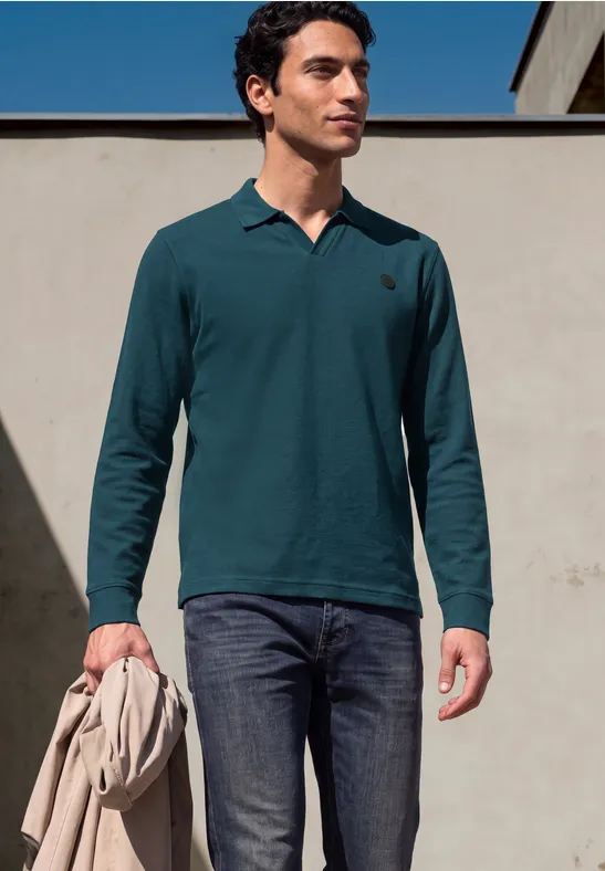 Ein Mann trägt ein petrolfarbenes Langarm-Poloshirt und dunkle Jeans und hält eine Jacke in der Hand.