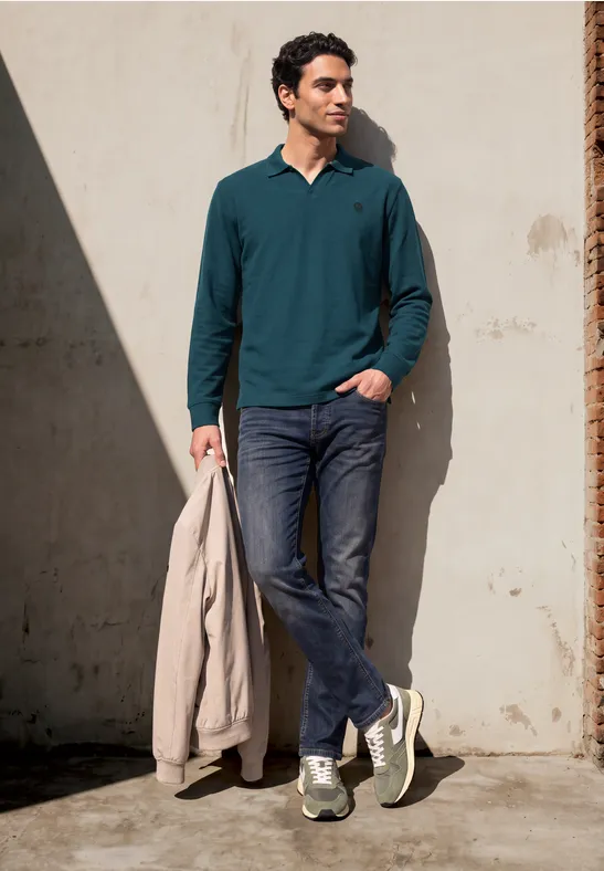 Ein Mann lehnt an einer Wand, trägt grünes Langarmshirt, Jeans, Sneaker und hält eine hellbeige Jacke.
