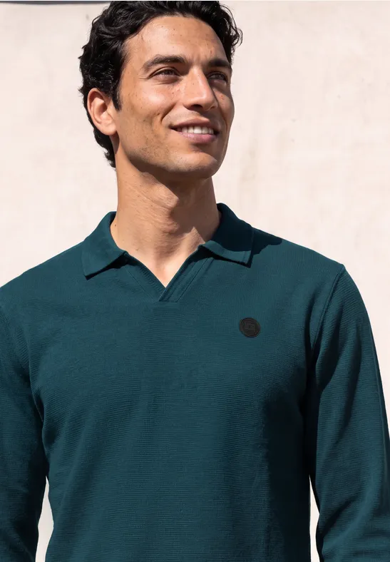 Ein lächelnder Mann in dunkelgrünem Langarm-Poloshirt vor hellem Hintergrund.